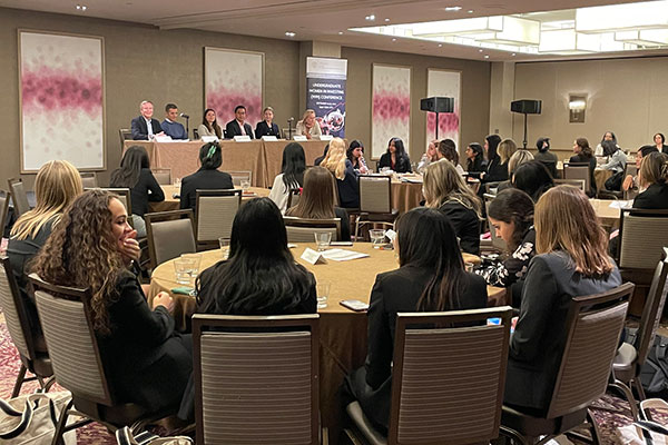 women in business attire seated at round tables facing a panel of speakers seated at a table on a stage.