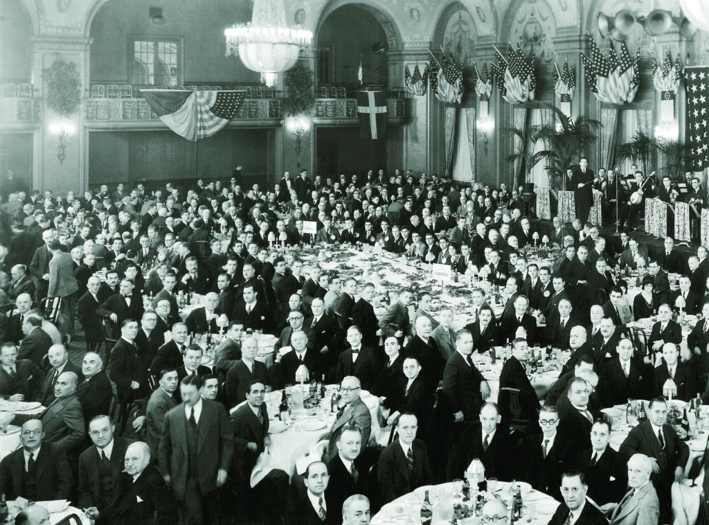 Black and white image of people in formal business attire in a grand banquet hall.