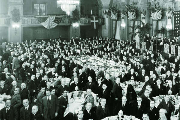 Black and white image of people in formal business attire in a grand banquet hall.