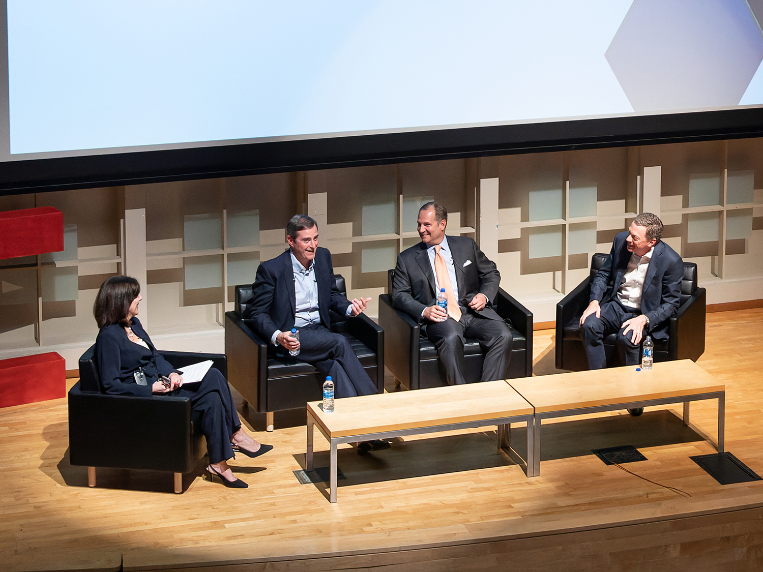 Four people sit in armchairs on a stage in front of a projector screen.