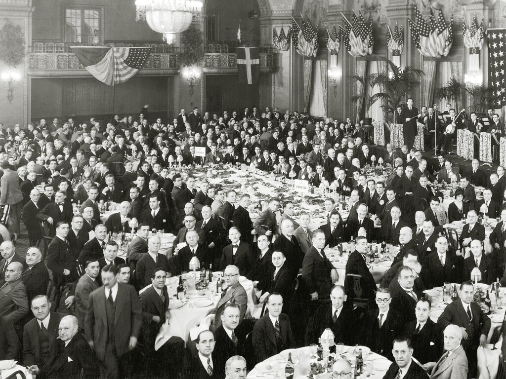 Black and white image of people in formal business attire in a grand banquet hall.
