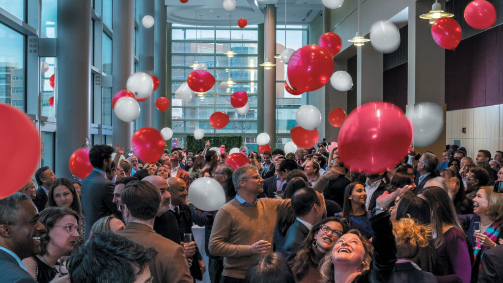 People stand and look up at red and white balloons falling from the above.
