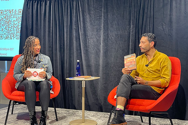 Illianna Acosta and Christopher Rivas seated, their chairs angled towards one another, looking at each other. rivas is holding up his book.