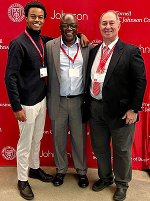 Photo of Paul Kavuma standing arm-in-arm with his son and Rob Canizares with a read Johnson Cornell banner in the background.