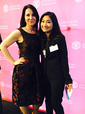 two women in business attire standing and smiling in front of a backdrop with the Johnson/Cornell University logo.
