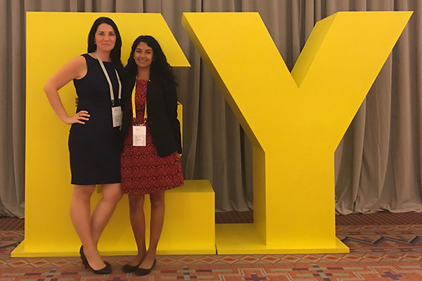 two women in business attire standing in front of a 3D EY logo.