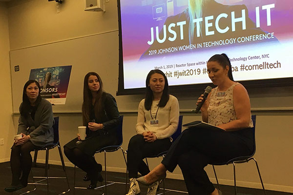 4 women, one speaking into a microphone, seated on high stools at the front of a conference room.