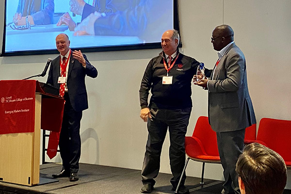 Andrew Karolyi standing at a podium and speaking with Rob Canizares and Paul Kavuam over on his right, Rob's hand on Paul's shoulder, all gesturing and smiling.