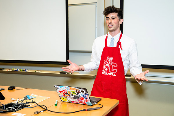 a young man in a red apron gesturing and delivering his pitch at the front of a room, laptop on the table in front of him.