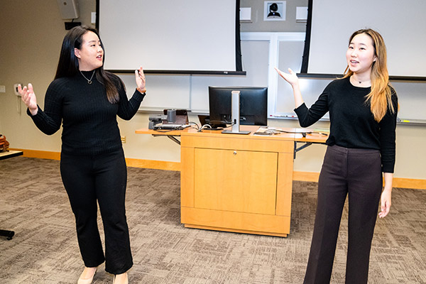 2 women at the front of a room gesturing as they deliver their presentation.