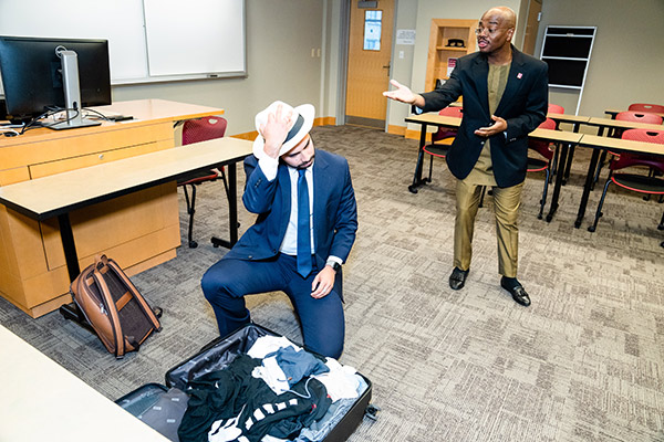 a young man in a business suit kneeling next to an open suitcase packed with clothes and putting on a hat and another young man gesturing towards him and speaking at the front of a room.