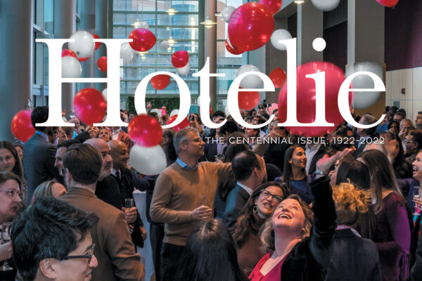 A large group of people celebrating the Hotelie Centennial indoors with red and white balloons floating in the air.