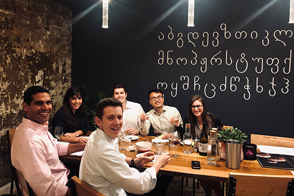 4 men and 2 women dressed in business attire sitting at a restaurant table and smiling. The Georgian alphabet is on the wall behind them.
