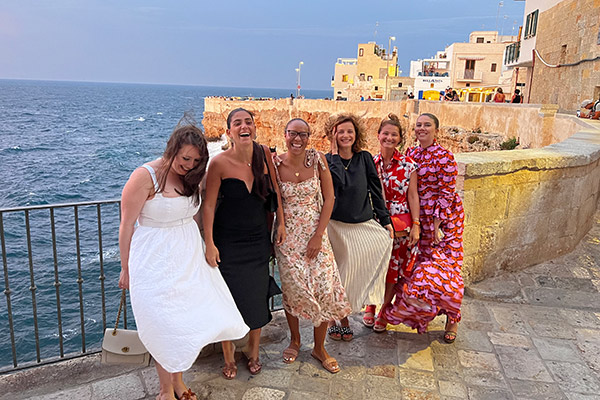 6 women in dresses, all laughing, on a windy day with a stone sea wall and the sea behind them.