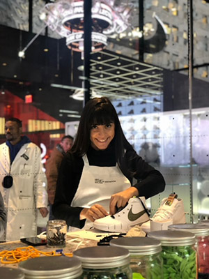 Mariam Kalandarishvili wearing an apron and standing at a factory table lacing a Nike shoe, looking up and smiling.