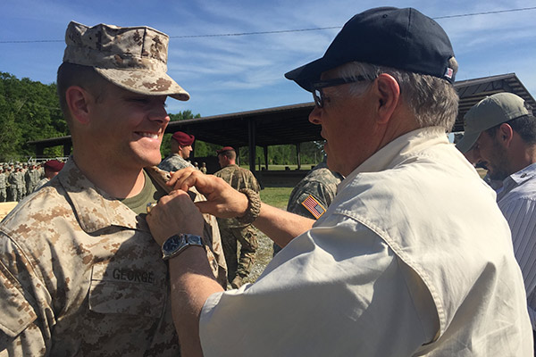 Patrick George wearing a camouflage Marine Corps uniform standing and smiling while his father pins his wings stud on his shirt. Other soldiers ae in the background.