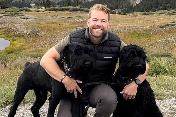 Patrick George, squatting, flanked by two large black dogs, his arms around he necks of each, in the middle of a green mossy field with evergreen trees in the background.