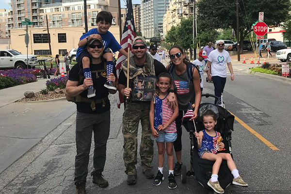 Patrick George standing on a city street with a young boy on his shoulders next to a man wearing camouflage and carrying an American flag. That man has his arm around a little girl standing in front of him and next to him are a woman and a young girl in a stroller. All the children and the woman have American flag motifs on their shirts. 