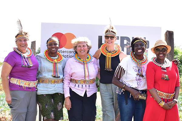 6 women, 3 Black and 3 white, standing next to each other, wearing Kenyan neck and head beaded adornments, and smiling.