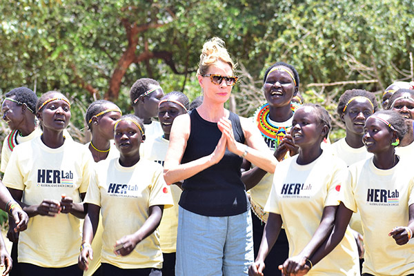 a white woman standing and clasping her hands together in appreciation, surrounded by teenage Kenyan girls who are singing and dancing.