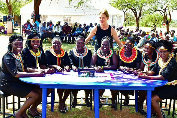 Linda Latsko Lockhart, arms outspread, standing behind a table of 8 young Kenyan women seated at a table, ready to play a card game.