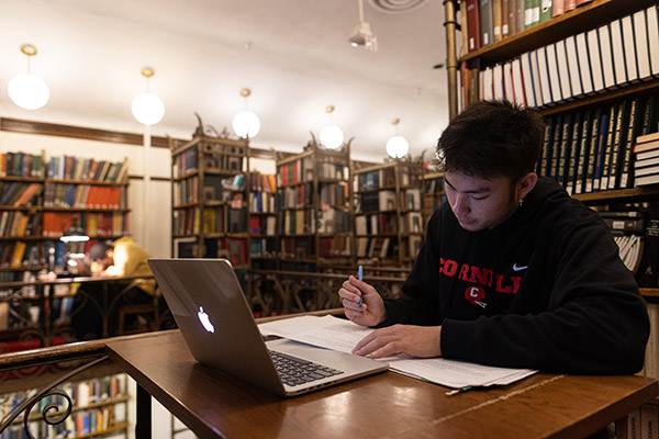 Jameson Wang seated at a desk with an open laptop and writing on one of the papers spread out in front of him, surrounded by bookshelves.