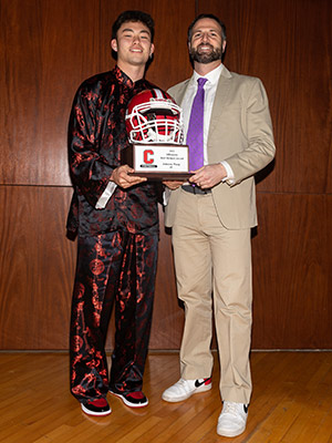 Jameson Wang wearing a red and black silk brocade shirt and pants and holding a helmet-shaped trophy standing next Coach Joe Villapiano, who is wearing a khaki suit.