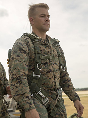 Patrick George standing in a field and wearing camouflage Marine Corps uniform and a parachute harness.