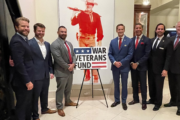 Seven men in business suits standing side by side around a "War Veterans Fund" sign,