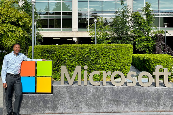 Kielyn Jarvis standing next to a large Microsoft sign in front of an office building landscaped with green shrubs and trees. 