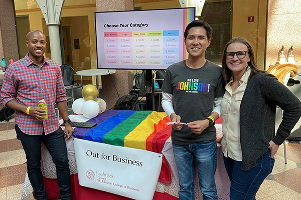 2 men and a woman smiling and standing on either side of a table draped with a rainbow banner and an Out for Business sign.