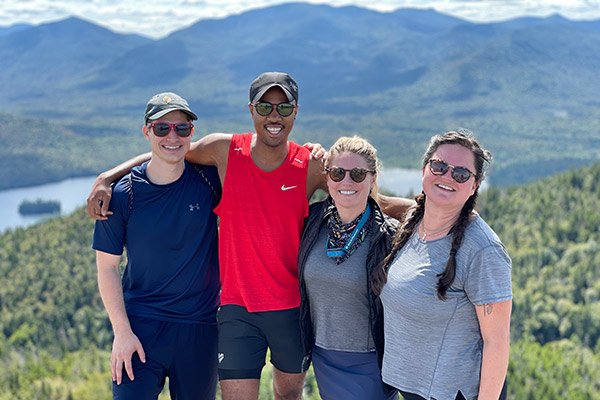 2 men and 2 women standing on top of a mountain on a sunny clear day with arms around each other’s shoulders. In the background are mountains, trees and a lake in the valley. 