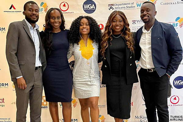 3 black women flanked by 2 black men, all dressed in business attire, standing in front of a backdrop with many different logos.