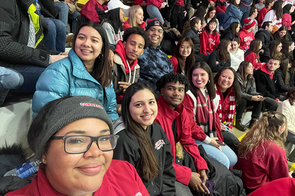 A selfie of Melanie Abreu with a large group of students sitting in bleachers with hats and coats on.