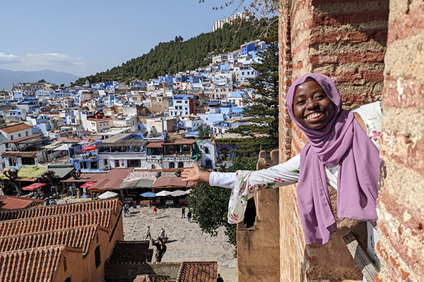 Mardiya Shardow leaning out of an opening in a brick building, smiling and gesturing to a mountainside village of blue and blue-roofed buildings.