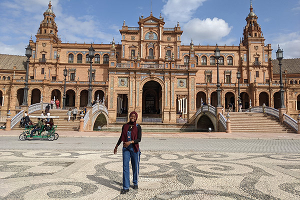 Mardiya Shardow standing on a broad plaza in front of a large, ornate building with curved staircases bridging a canal leading to it.