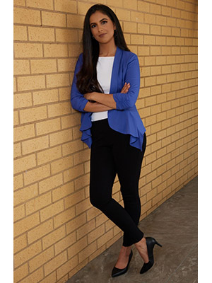 Nikita Gossain in a blue jacket and black pants and high heels leaning again a brick wall and smiling.