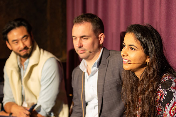 A close-up of 2 men and 1 woman sitting next to each other, wearing microphone headsets. The woman, Nikita Gossain, is speaking and smiling.