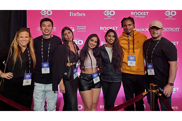 A group of 4 women and 3 men standing arm-in-arm and smiling in front of a Forbes 30 Under 30 backdrop.