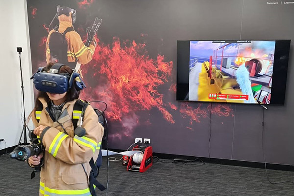 A woman standing in a carpeted room, wearing a virtual reality headset, and holding the large nozzle of a fire hose with both hands.