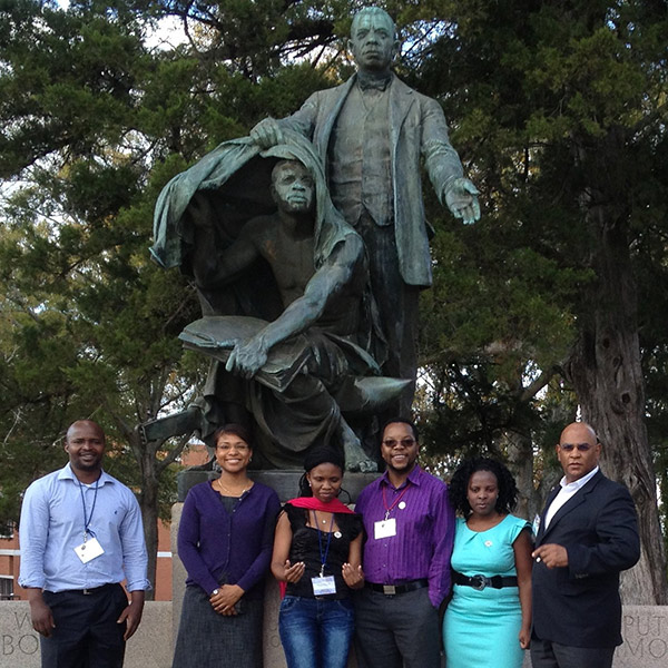 3 Black women and 3 Black men standing in front of a statue of Booker T. Washington lifting the cover off a man crouched below him. 