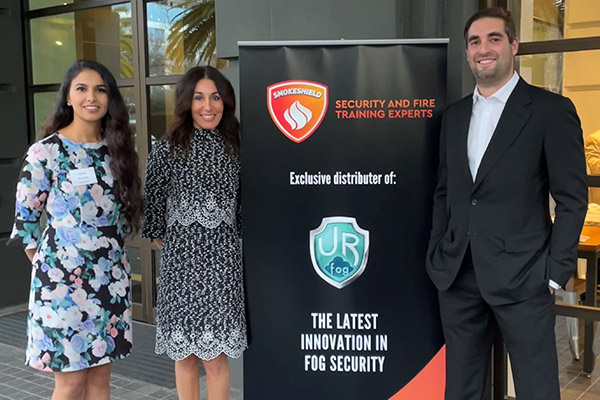 2 women and a man standing on the glass-windowed entryway to a building next to a banner announcing Smokeshield as the exclusive distributor of Ur Fog.