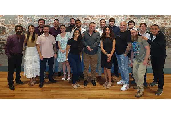 A group photo of 18 men and women standing in front of a brick wall, laughing and smiling. 