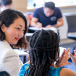 2 women sitting in a classroom and speaking with one another with other students seated and writing in the background.
