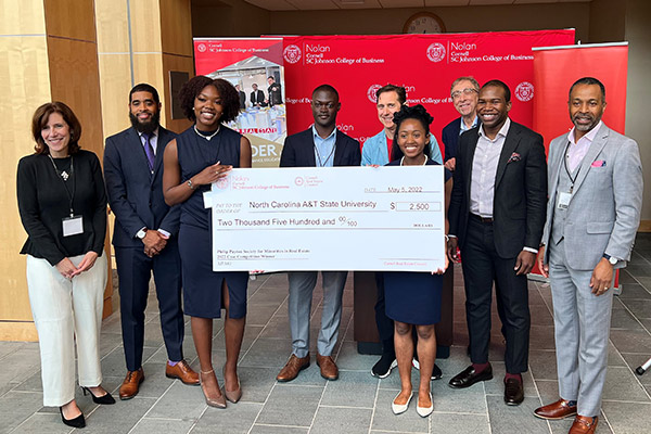 2 young Black women, dressed in business attire, are holding a novelty check for $2,500 made out to North Carolina A&T State University. Surrounding them are 7 other people: 1 woman and 6 men.