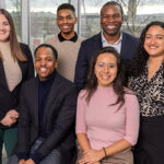 3 men and 3 women dressed in business casual posing in front of a window and smiling.