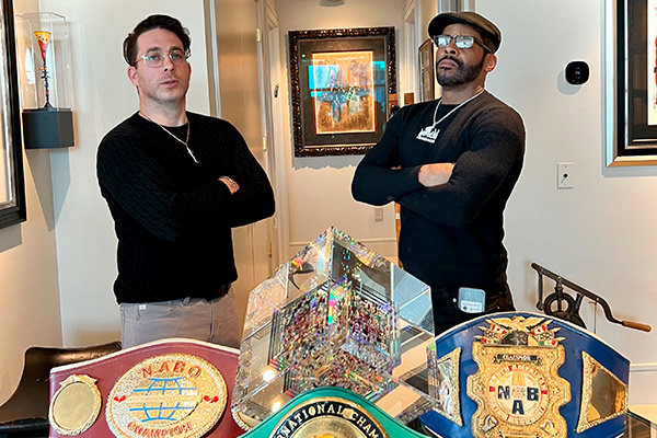 two men standing with arms folder and heads held high in front of a table of trophies.