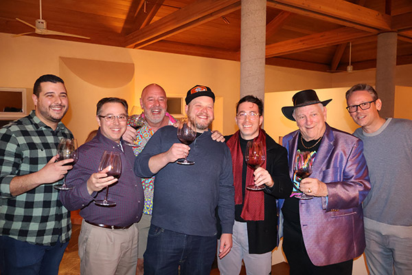 7 men standing, smiling, and holding up glasses of wine.