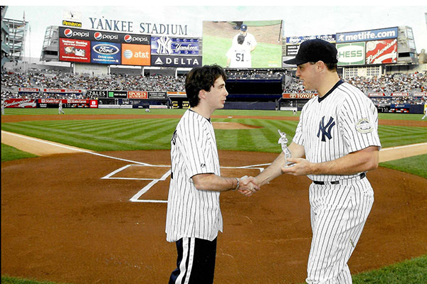 A boy and a man, both in Yankees baseball uniforms, shaking hands on the ball field at Yankee stadium.
