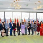 a group of 21 men and women standing in front of many flags from different nations and large windows with a large building behind them.